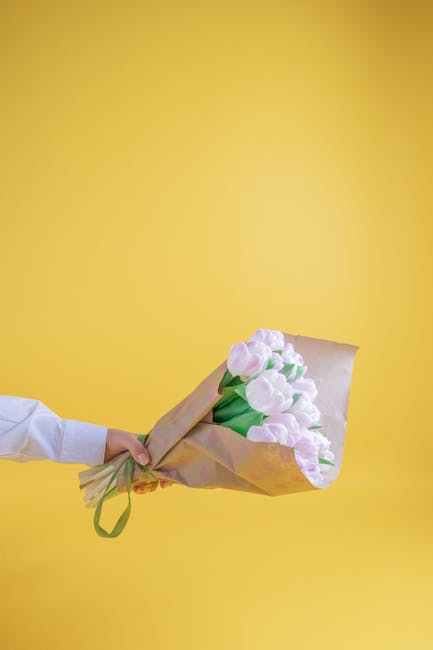 A hand extending from the left holds a bouquet of fresh, pale pink tulips wrapped in light brown paper with green ribbon, positioned against a solid bright yellow background. The tulips have smooth, s