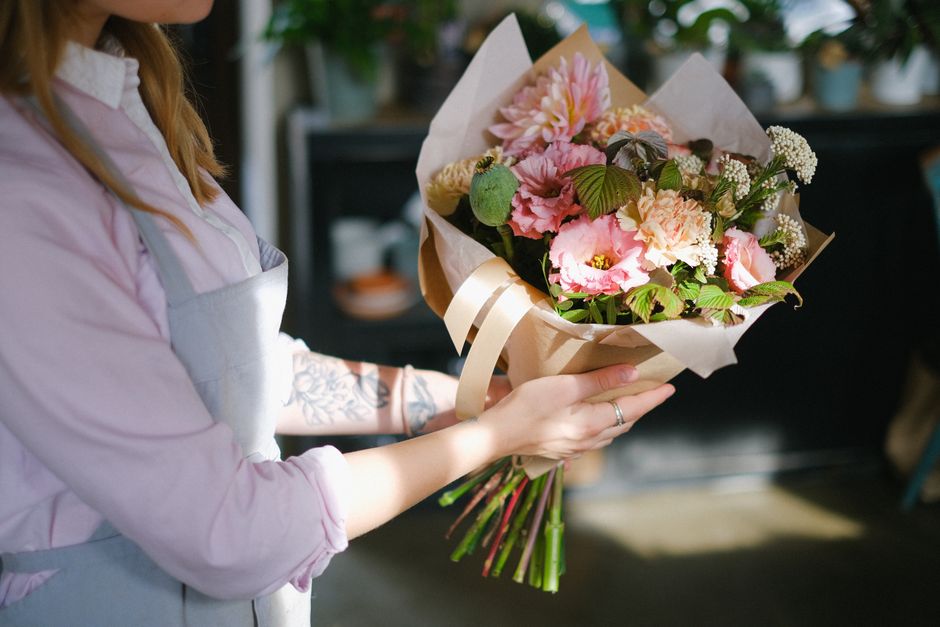 A person wearing a light pink shirt and a grey apron is holding a bouquet of fresh flowers in a floral arrangement, featuring pink snapdragons, pale peach carnations, white baby's breath, and green fo