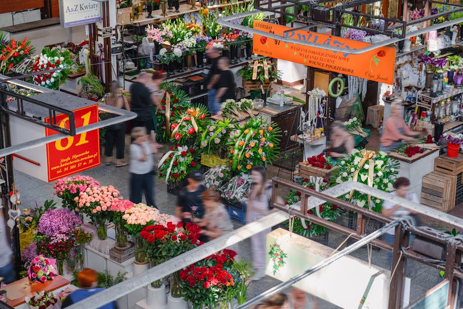 A bustling indoor flower market showcasing a vibrant array of floral arrangements and bouquets. In the foreground, there are pots and vases filled with pink, red, and purple chrysanthemums and roses,