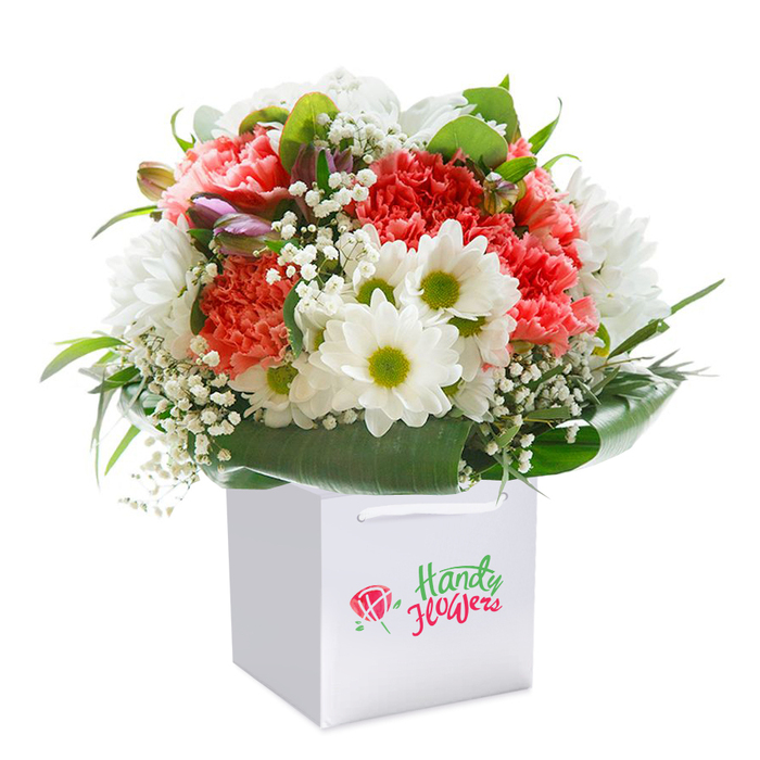 Standing tall within a clean, square white gift bag, this vibrant bouquet is a showcase of Greenhithe's floristry expertise. Softer shades of coral and peach mingle seamlessly with lush red carnations, their ruffled petals brimming with texture and visual warmth. Sprightly white daisies, crowned by green-yellow centers, interject splashes of joyous contrast, reminiscent of the wildflower verges near Ingress Abbey. Baby's breath, with its fine, misty clusters, gently weaves a feeling of serenity and airiness throughout, while on the arrangement's left, a few unopened buds tinged with purple hint at life's unfolding possibilities-much like new spring growth along Knockhall Chase. Deep green, glossy foliage frames the bouquet in a graceful arc, offering both structural support and a verdant backdrop. The white gift bag, finished with smooth ribbon handles, speaks to the convenience and care of local Greenhithe flower delivery, ensuring every bouquet arrives immaculate for birthdays, anniversaries, or thoughtful tokens. Clear local branding on the bag subtly reinforces the trust and attention to quality customers expect. The bouquet's abundant, rounded form and harmonious blend of colours and textures reflect the hand of a true local florist, ready to bring a fresh sense of celebration and connection to any Greenhithe address.