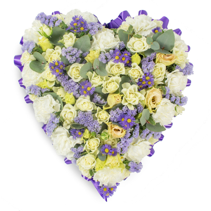 Presented against a pristine white backdrop, this heart-shaped memorial arrangement is a study in soft contrasts and tender craftsmanship, the sort of composition a florist might prepare for a chapel service on the Thames foreshore. The core is a dense collection of cream and pale yellow roses, ranging from button-like buds to fuller, gently opening blooms that give the heart depth and a buttery, velvety texture. Interspersed among them are ruffled white carnations that provide a classic, comforting fullness; small pale yellow flowers lift the warmth subtly, while sparing clusters of lavender statice and vivid purple aster-like blooms with yellow centres add delicate, jewel-like accents that animate the surface. Rounded eucalyptus leaves in blue-green tones are placed with intention to create visual pauses, their matte sheen and fresh scent balancing the sweetness of the roses. Encircling the entire heart is a continuous band of rich purple satin ribbon, looped and arranged to trace the outline and confer a refined finish. The overall impression is peaceful and consoling - dignified for wakes, funeral services, or graveside memorials in Greenhithe, near the station or riverside paths - a tender expression of enduring love and respect. The arrangement's textures, soft colour palette and subtle fragrance together form a comforting presence for those remembering a loved one in our local community.