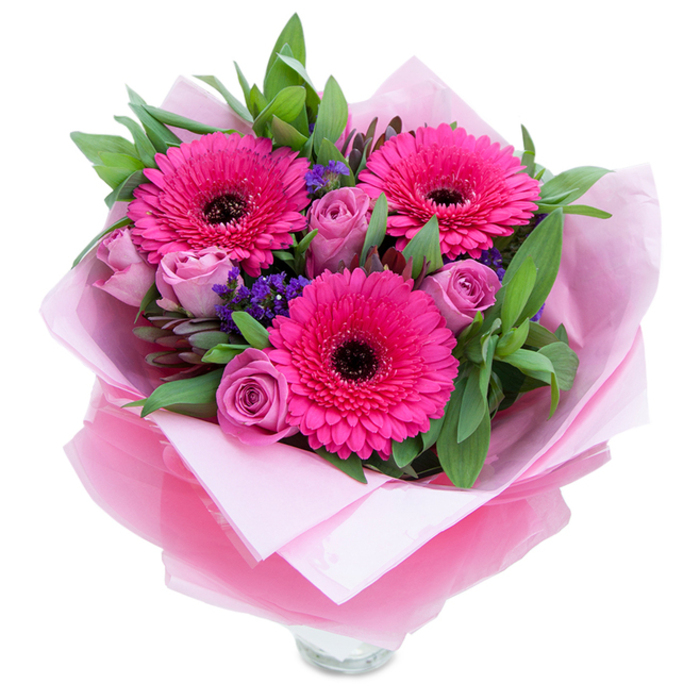 There is a playful elegance to this bouquet that I describe as a florist familiar with Greenhithe's gentle rhythms - the bright intensity of fuchsia set against softer, dusty rose notes. Three magnificent fuchsia Gerbera daisies command attention with their abundant radial petals and dark, almost black centres that provide a bold punctuation in the centre of the arrangement. Surrounding them, roses in a muted lavender-pink unfurl in layered, velvety spirals; their buds are fresh and compact, promising a slow reveal of fragrance that is tender and reminiscent of garden roses on a cool morning. Interspersed among the principal blooms are fine stems of purple statice, their tiny clustered florets creating a lightly airy texture that seems to float and catch the eye between larger shapes. A healthy foundation of greenery - broad, glossy leaves like salal and hints of eucalyptus - frames the flowers with a clean, verdant outline and offers a crisp, green aroma when handled. For presentation the bouquet is cocooned in soft pale pink tissue, with an outer wrap of translucent fuchsia-pink cellophane that shimmers slightly under soft shop lighting, folded with careful precision around the stems to form a pleasing silhouette. The clear glass vase rim is visible at the base, suggesting the bouquet is ready to be set on a café table in Greenhithe or placed at the centre of a windowsill that looks toward the Thames. The overall effect is warm, celebratory and comforting - fitting for a birthday, anniversary, or a thoughtful 'just because' gift for a neighbour or friend in the local community near Ingress Park.