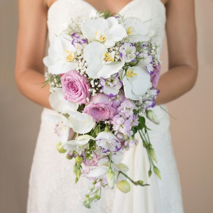 This refined, close-up image captures the moment a bride from Greenhithe stands poised, her arms cupping a lavish cascading bouquet that spills gracefully downward, the soft sunlight echoing the gentle glow of the Thames on a clear afternoon. Dominating the bouquet's lush composition are large, pure white phalaenopsis orchids, their satin petals wide and arching, revealing sunny yellow and faint purple details at their center. Among them, generous heads of blush-pink garden roses nestle, their petals unfolding in layers that evoke romance and gentle tradition. Lavender stock and delicate light purple lisianthus flowers mingle through the design, adding movement, texture, and a whisper of wild elegance-like the varied flora along Greenhithe's coastal trails. Scattered white eustoma, often edged with richer purple, punctuate the display, while baby's breath (Gypsophila) clouds the bouquet in soft, ethereal fullness. Minimal yet purposeful greenery creates structure and balance without overwhelming the gentle colour story. From the strapless bodice and lace-detailed upper skirt of the bride's gown, to the sense of careful arrangement and natural movement, every aspect draws admiration. This bouquet, a picture of Greenhithe's signature style and sophistication, would make a centerpiece at any celebration, perfectly reflecting the skill and artistry of Flower Delivery Greenhithe's talented florists.