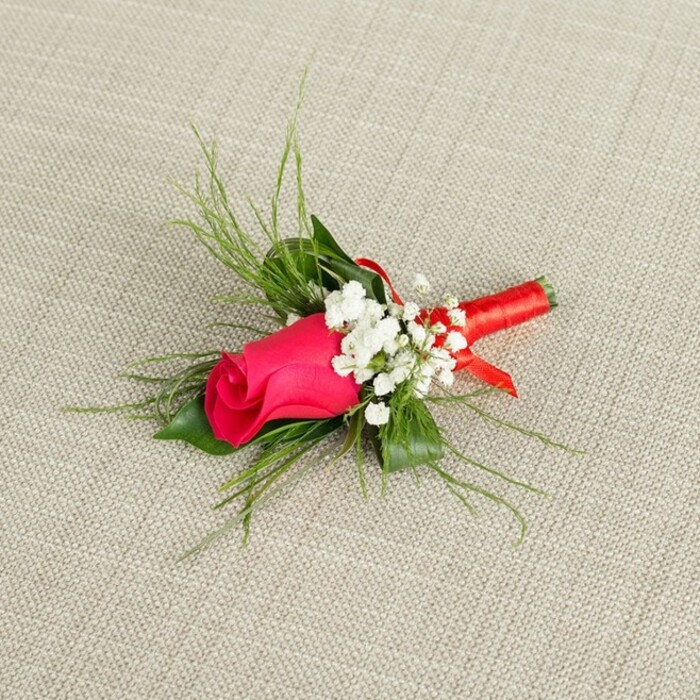 Set on a softly textured, light beige fabric that looks like gently woven linen, this boutonnière is captured at a diagonal angle, as if just placed down for a moment in a quiet corner of a Greenhithe dressing room before a ceremony. At the centre, a single rosebud in a striking fuchsia or deep magenta pink draws the eye, its petals tightly coiled and smooth, with that velvety sheen that only a fresh rose can carry. The bud is not yet open, giving it a sense of anticipation and youthful elegance, ideal for a groom, groomsman, or prom guest about to step into a special occasion. Surrounding the rose, fine clusters of white baby's breath scatter like tiny stars, their numerous miniature blossoms creating a soft, airy cloud that lightens the overall design and offers a delicate counterpoint to the strong, saturated hue of the central bloom. Behind and around these flowers, wisps of bright green, feathery foliage spread out in graceful arcs, giving the piece movement and an almost hand-drawn, sketch-like quality, as if nature has been gently coaxed into perfect form. Closer to the base, a few darker green leaves ground the arrangement, adding depth and structure without drawing focus away from the rose. The stems are skillfully aligned and bound in a narrow band of rich red satin ribbon, wrapped in smooth, even coils, with a subtle knot or small bow detail that suggests the expert hands of a local florist who understands how these pieces must sit neatly against a lapel. The ribbon's sheen catches the light, echoing the soft highlights on the rose petals. The lighting is bright yet gentle, with no harsh shadows, allowing every edge, curve, and thread of foliage to be seen clearly. The composition feels thoughtfully balanced and refined, like a traditional boutonnière crafted for weddings along the Thames or formal celebrations in and around Greenhithe, where understated elegance and heartfelt detail are cherished.