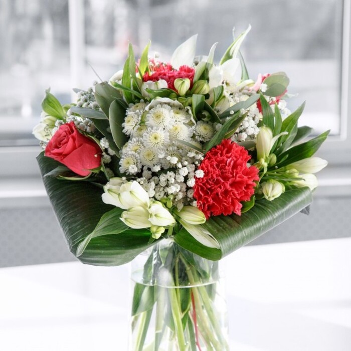 A refined bouquet of red and white flowers rises from a crystal-clear cylindrical vase, the stems inside aligned with the precision of a florist who knows every angle matters. At first glance, the deep red roses command attention, their velvety petals swirling outward in perfectly formed spirals that evoke timeless romance. Just below them, a large crimson carnation, densely ruffled and rich in texture, creates a tactile focal point, its frilled edges catching the soft light like fabric. These bold reds are balanced by an array of white and creamy blooms, including elegant white alstroemeria whose slim petals and fine inner markings add subtle intricacy. Several white lily buds are still tightly closed, standing tall among the open flowers, suggesting that the bouquet will continue to evolve over the coming days, much like the changing light along the riverfront in Greenhithe. Throughout the composition, airy sprays of white gypsophila fill the spaces between the larger blossoms, adding a soft, cloud-like veil that gives the arrangement a sense of fullness without heaviness. Around the base, glossy dark green leaves are carefully curled and wrapped to frame the bouquet, forming a structured, almost architectural collar. Additional foliage in varied shades of green, including eucalyptus-style leaves and slender pointed stems, weaves through the design, introducing movement and a natural, garden-fresh character. The arrangement is set on a pristine white surface, with a gently blurred, luminous background that suggests daylight streaming into a Greenhithe home or office. The overall atmosphere is both celebratory and calm, as if designed for a special table on Stone Place Road or a thoughtful gift delivered to a loved one nearby, perfect for anniversaries, Mother's Day, or simply to say I care with quiet elegance.