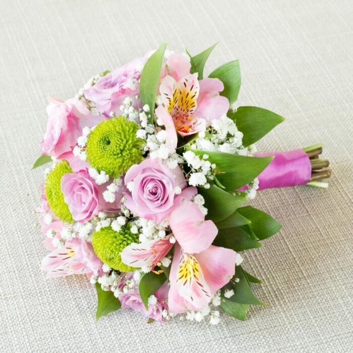 A close-up view reveals a sumptuous, hand-tied bouquet resting diagonally across an understated, textured beige surface reminiscent of a classic table in Greenhithe's cosy cafes. The heart of the bouquet features pastel pink and mauve roses, each blossom carefully unfurled to display intricate spiral centres and a silken sheen, offering both visual and tactile delight. Set against them, lime-green button chrysanthemums form bright, perfect spheres, providing lively contrast and a modern touch. Interspersed pink alstroemeria unfurl softly, revealing slender petals adorned with delicate yellow and russet speckles, nodding gracefully as if caught in a gentle breeze near Ingress Abbey. Wisps of snowy white baby's breath appear throughout, creating a sense of airiness and unity, as though the bouquet is kissed by a morning fog. The lush, dark green leaves peeking beneath the blossoms lend the piece both structural depth and freshness, evoking Greenhithe's riverside greenery. All stems are cleanly gathered and bound with a vivid, magenta satin ribbon, wrapped tightly but artfully, leaving the cut stems visible, hinting at the arrangement's farm-to-vase authenticity. Soft, even light bathes the bouquet, highlighting subtle colour gradients and textures. This meticulous composition by Flower Delivery Greenhithe exudes romance, happiness, and the optimism of spring, making it a perfect choice for bridal parties, anniversaries, or as a radiant gift delivered to homes throughout the Greenhithe area.