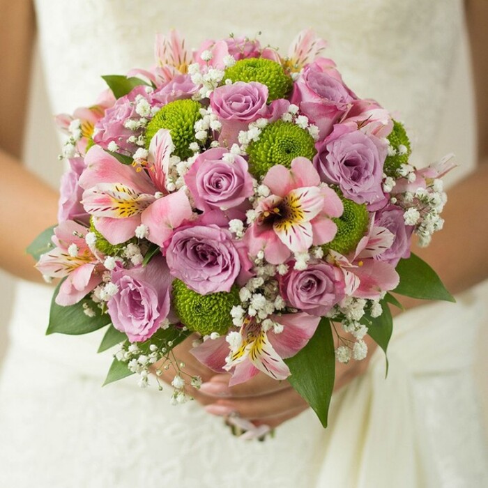 Nestled against the softly textured lace of a Greenhithe wedding dress, this exquisite bridal bouquet tells a story of love, artistry, and attention to detail. The spherical design overflows with pastel roses-blush pink and muted lavender-whose petals glow with a fresh softness, some kissed by the glimmer of dew. Vibrant green button chrysanthemums, perfectly round and bold, inject a contemporary zest into the arrangement, reminiscent of the lush local scenery near the Thames. Pink alstroemeria lilies, their petals brushed with golden yellow and veined in deep magenta, weave through the bouquet, adding an exotic and sophisticated flourish. Wisps of airy white gypsophila, or baby's breath, infuse the bouquet with lightness and volume, uniting the larger flowers in gentle harmony. At the base and edges, glossy dark green leaves frame the scene, grounding the bouquet with natural structure. The bride's hands, one adorned with a simple gold band, cradle the stems tenderly, embodying hope and excitement before her special day in Greenhithe's charming venues. This bouquet is a botanical love letter-a vibrant accent for celebrations set amidst Greenhithe's riverside beauty, arranged by expert local florists.