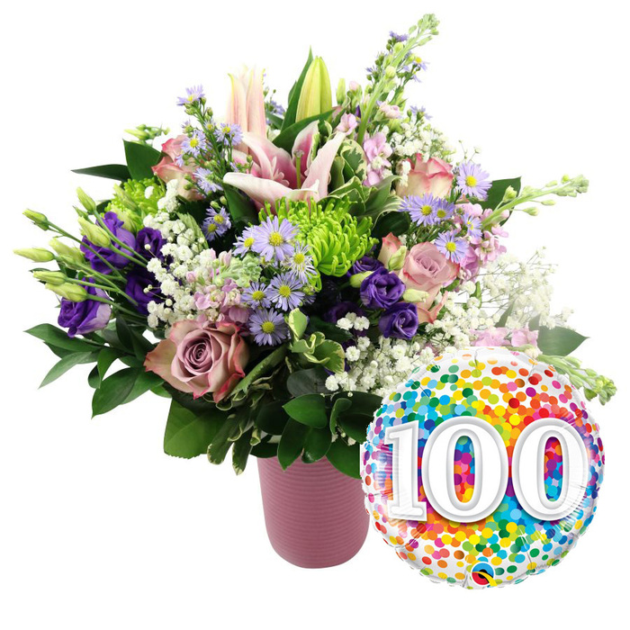 Captured against a spotless white background, this exuberant floral arrangement and milestone balloon combination looks like it has just been prepared for a 100th celebration in Greenhithe. The flowers spill generously from a tall, cylindrical vase finished in a soft dusty-pink hue, its matte, ribbed surface adding a modern, tactile quality. Dominating the arrangement are several light pink Oriental lilies: some blossoms are fully open, revealing broad star-shaped petals that curve gently backwards, while others remain in slender bud form, adding height and an air of anticipation. Nestled around the lilies are roses in complementing shades of soft pink, mauve, and dusty rose, their layered petals unfolding in gentle spirals that evoke classic romance and long-lasting affection. Threads of deep purple lisianthus wind between the roses and lilies, their velvety, cup-like blooms bringing depth and richness. Scattered amid the larger flowers are sprays of dainty lavender asters with tiny yellow centres, echoing the carefree feel of wildflowers one might spot on a walk near the Greenhithe waterfront. Compact, lime green button chrysanthemums dot the composition with playful bursts of colour and tightly packed texture, standing out against the pastel tones. Airy clusters of white baby's breath float through the arrangement like fine lace, adding softness and volume. A surround of dark green foliage frames the entire bouquet, enhancing the colours and creating a lush, full silhouette. To the lower right, slightly overlapping the base of the flowers, a round foil balloon gleams under the light. Its surface is covered in a lively pattern of multi-coloured confetti dots in red, orange, yellow, green, blue, and purple, while bold white numerals reading 100 stand proudly in the centre with a subtle shadowed effect. The overall impression is one of joyful respect and heartfelt celebration, perfectly suited for marking a 100th birthday or anniversary in Greenhithe, from cosy homes near the station to family gatherings overlooking the river.