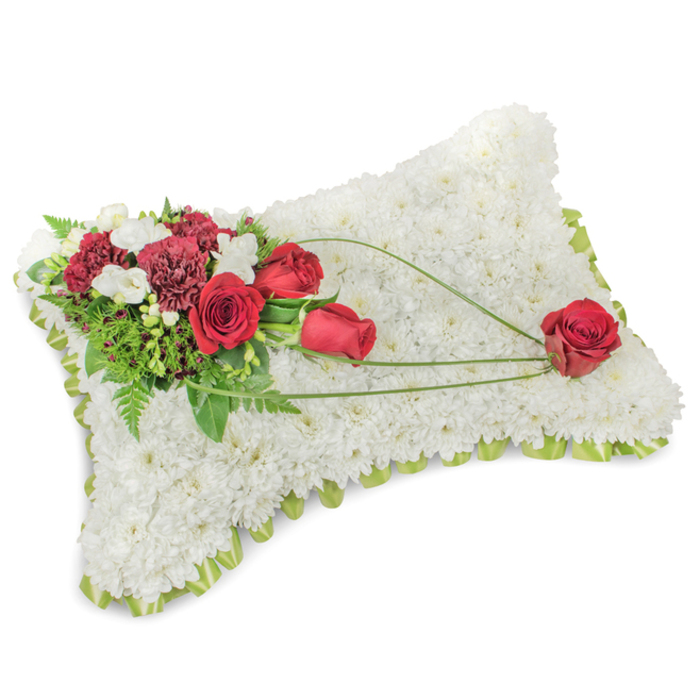 A beautiful, poignant memorial pillow captured on a plain white backdrop, where every petal and curve is visible as if in gentle morning light. The rectangular cushion is meticulously constructed from hundreds of small, immaculate white chrysanthemums, pressed close to create a sumptuous, tactile surface that reads as both comforting and pure. A subtle rim of pale lime-green ribbon and glimpses of delicate foliage edge the pillow, providing a soft frame that lifts the blooms and adds a faint, fresh colour contrast. On the upper left corner rests a lavish spray of deep red flowers - roses of satin-like texture and darker burgundy carnations - their shapes layered and dramatic against the white ground. Small white accent flowers, likely freesias or spray mums, nestle among the reds, giving points of brightness and a whisper of fragrance. Supporting greenery, from ferny fronds to denser glossy leaves, forms a verdant backdrop, increasing depth and richness. From this main cluster, sinuous lines of light green bear-grass or similar foliage extend diagonally across the cushion toward the lower right, creating a graceful visual path that leads to a single, perfectly formed red rose positioned as the arrangement's quiet focal point. The composition balances precision and natural flow; the textures shift from the chrysanthemum's soft fleece-like surface to the dense velvet of rose petals, while the overall scent remains restrained and respectful. This dignified piece would be appropriate for funerals, memorial services, or private remembrance moments in Greenhithe, near the Thames riverside, delivering a message of solace, honour, and enduring affection.
