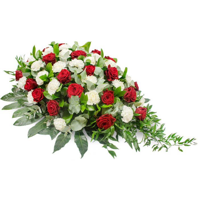 Photographed on a stark white background, this sophisticated funeral floral spray takes on a sculptural presence, its elongated shape and carefully balanced colours clearly defined. The arrangement is horizontal and asymmetrical, with its fullest, richest cluster of blooms gathered to the top left. From there, the flowers and greenery gradually taper down and outward toward the bottom right, forming a graceful teardrop or crescent that suggests movement and gentle release. The main blooms are an opulent mix of deep red and pure white flowers. The red roses, with their layered, velvety petals, are scattered throughout the spray, creating points of emotional intensity that symbolise love, devotion, and a grief that is deeply felt. Between and around them, generous white roses and white carnations provide contrast in both colour and texture; their smooth or soft, fringed petals offer an impression of innocence, reverence, and quiet solace. Nestled beneath the flowers lies a diverse selection of foliage. Broad, muted sage-green leaves give structure and a velvety look, while darker green leaves deepen the background. Delicate, fern-like fronds and finer trailing stems extend beyond the main outline, especially on the right-hand side, lending the design a soft, cascading rhythm. The overall palette-crimson, white, and layered greens-feels both traditional and contemporary, suitable for a range of memorial settings. For families in Greenhithe, a piece like this could be seen resting with dignity at a local crematorium or chapel, perhaps after being created by Flower Delivery Greenhithe for a service near the riverfront, offering a gentle yet eloquent floral expression of respect and farewell.