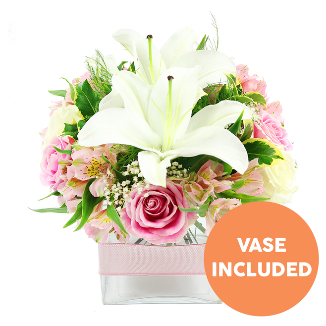Against a clean, luminous white background, a lovingly crafted floral arrangement takes centre stage, exuding a mix of elegance and warmth. Two prominent white Oriental lilies anchor the design, their large petals opening generously and curling back to reveal striking dark reddish-brown stamens that add drama and focal interest. Gathered closely around them are numerous soft pink roses, their hues ranging from blush to slightly deeper rose, some still in tightly curled buds and others fully open, displaying lush, multi-layered whorls of petals that suggest romance and heartfelt emotion. Slender stems of alstroemeria thread through the bouquet, their pink and white petals sometimes tinged with peach and decorated with subtle yellow markings, bringing texture and a relaxed, garden-grown charm, much like the flowerbeds you might see in local Greenhithe gardens or near the riverbank. Between the larger blooms, tiny clusters of white baby's breath form light, airy tufts that soften the composition and give it a gentle, cloud-like quality. A rich mix of greenery holds everything together: broad, glossy leaves provide structure and sheen, while finer, feathery foliage introduces softness and depth, shaping the bouquet into a pleasing, slightly domed form. The flowers sit securely in a square, clear glass vase that reveals the green stems beneath the water, lending a clean, contemporary look. Around the middle of the vase, a pale pink satin ribbon runs horizontally, its smooth texture catching the light and echoing the pink tones of the roses. In the bottom right corner, a vivid orange circular badge stands out clearly, the words VASE INCLUDED printed in bold white capital letters to highlight that this is a complete, ready-to-place arrangement. The overall mood is serene yet celebratory, ideal for brightening a Greenhithe apartment, marking an anniversary, or sending a gentle message of caring with local flower delivery.